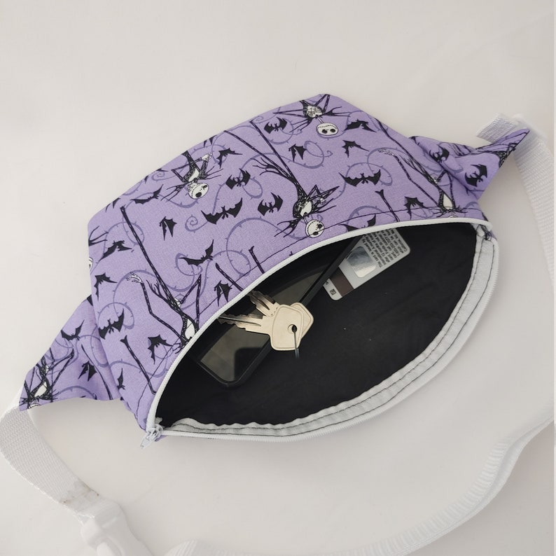 Może przedstawiać: Fioletowa nerka z czarno-białym nadrukiem Jacka Skellingtona i nietoperzy. Nerka jest otwarta i pokazuje czarne wnętrze z telefonem i kluczami.