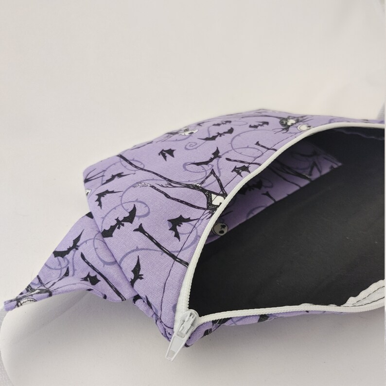 Może przedstawiać: Fioletowa kosmetyczka zapinana na zamek błyskawiczny z białym zamkiem i czarno-białym nadrukiem nietoperzy i postaci szkieletu. Kosmetyczka jest podszyta czarną tkaniną.