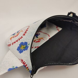 Puede incluir: Bolsa blanca con cremallera y forro negro. La bolsa tiene un fondo blanco con un estampado floral azul y amarillo y un texto rojo que dice "Friends F.E.A.N.S. Ohana".