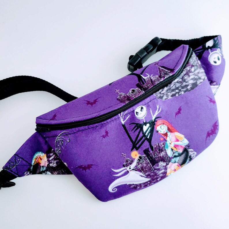 Może przedstawiać: Fioletowa nerka z czarnym zamkiem błyskawicznym i regulowanym paskiem. Tkanina ma nadruk Jacka Skellingtona i Sally z filmu Koszmar przed świętami.