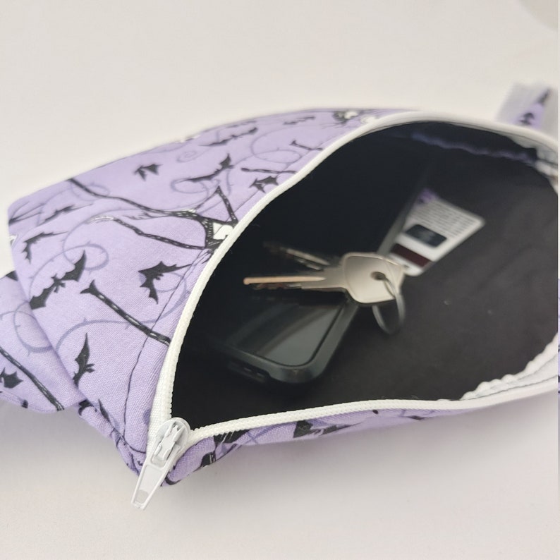 Może przedstawiać: Fioletowa kosmetyczka zapinana na zamek błyskawiczny z czarno-białym wzorem nietoperzy. Kosmetyczka jest otwarta i widać w niej czarny telefon kom&oacute;rkowy i klucze.