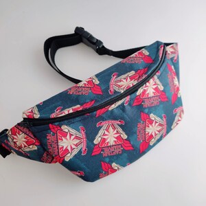 May include: A black fanny pack with a zipper closure. The fabric is a dark blue with a repeating pattern of red and gold Captain Marvel logos.