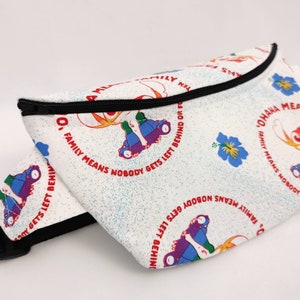 Puede incluir: Ri&ntilde;onera blanca con cremallera negra y un patr&oacute;n repetido de patines de colores y flores. El texto "Ohana Family Means Nobody Gets Left Behind Or Forgotten" est&aacute; impreso en rojo en la bolsa.