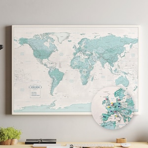 May include: A light blue world map with a white background and black text. The map is framed and hanging on a white wall above a desk. A smaller, circular map of Europe is on the desk with colorful pins marking locations.