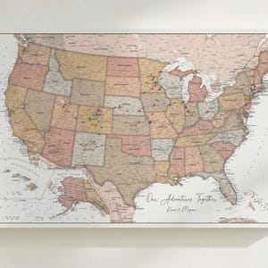 May include: A framed map of the United States with a vintage aesthetic. The map is divided into states with various colors and includes pins marking locations. The text "Our Adventures Together" is written in cursive, along with two names.