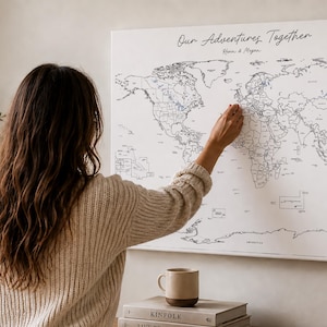 May include: A canvas world map with black outlines and the text "Our Adventures Together" is displayed on a wall. A person is pointing at the map. A mug, books, and a wooden bowl are on a wooden surface.