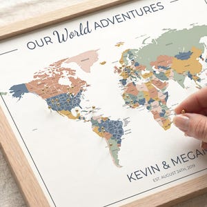 Puede incluir: Un mapa del mundo enmarcado con alfileres que marcan ubicaciones. El mapa está en colores apagados y tiene el texto "OUR WORLD ADVENTURES" en la parte superior. Los nombres "KEVIN & MEGAN" y la fecha "EST. AUGUST 24TH, 2019" están impresos en la parte inferior.