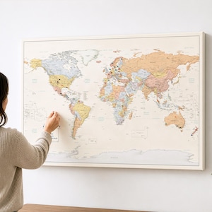 May include: A framed world map with push pins marking locations. The map is in a neutral colour palette and is mounted on a white wall. A person is pointing at the map. The map is a decorative piece.