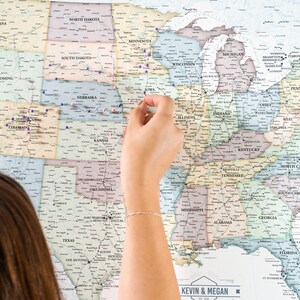 May include: A vintage-style map of the United States with push pins marking locations. The map is on a wall and a person's hand is placing a pin on the map. The map has the text "Kevin & Megan Est. 2019" and "Past Adventures" and "Yet to Explore" at the bottom.