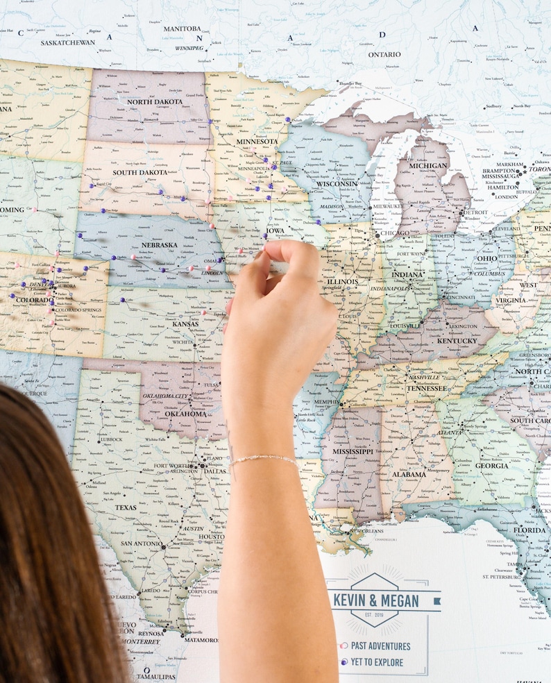 May include: A vintage-style map of the United States with push pins marking locations. The map is on a wall and a person's hand is placing a pin on the map. The map has the text "Kevin & Megan Est. 2019" and "Past Adventures" and "Yet to Explore" at the bottom.