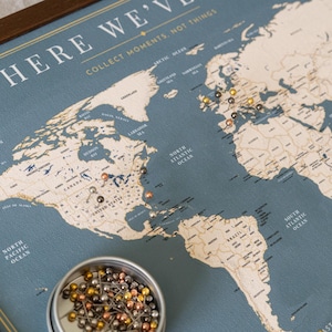 May include: A world map with a blue background and a wooden frame. The map is marked with pins of various colors, indicating locations. The text "WHERE WE'VE" and "COLLECT MOMENTS, NOT THINGS" is visible. A small container of pins sits in the corner.