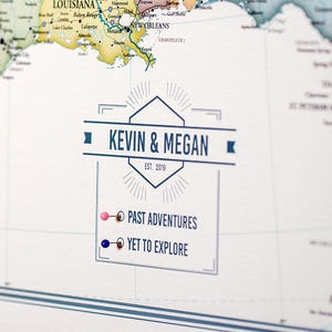 May include: A white map with a blue and white banner that says "KEVIN & MEGAN EST. 2019". There are two push pins, one pink and one blue, with text that says "PAST ADVENTURES" and "YET TO EXPLORE".