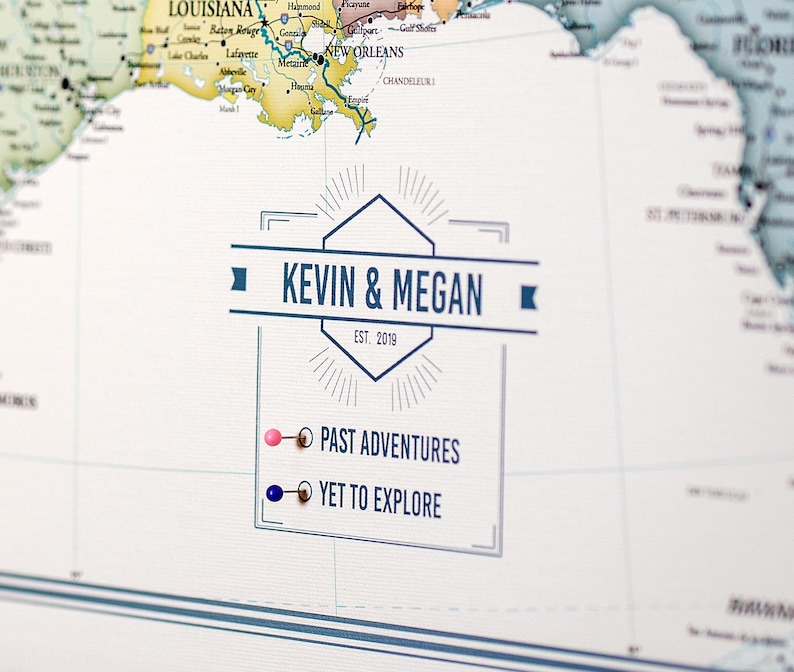 May include: A white map with a blue and white banner that says "KEVIN & MEGAN EST. 2019". There are two push pins, one pink and one blue, with text that says "PAST ADVENTURES" and "YET TO EXPLORE".