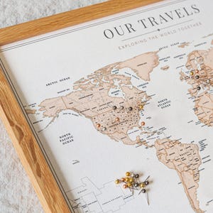 May include: A framed world map with the text "OUR TRAVELS EXPLORING THE WORLD TOGETHER". The map is in shades of brown and beige, with pins marking locations. The frame is made of light-colored wood. A collection of pins is scattered near the bottom.