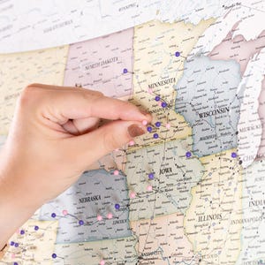 May include: A close-up of a hand placing a purple push pin on a map of the United States. The map is covered in purple and pink push pins, indicating locations across the country.