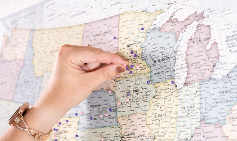 May include: A close-up of a hand placing a purple push pin on a map of the United States. The map is covered in purple and pink push pins, indicating locations across the country.