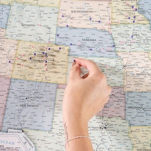 May include: A close-up of a map of the United States with push pins marking various locations. A hand is placing a push pin on the map.