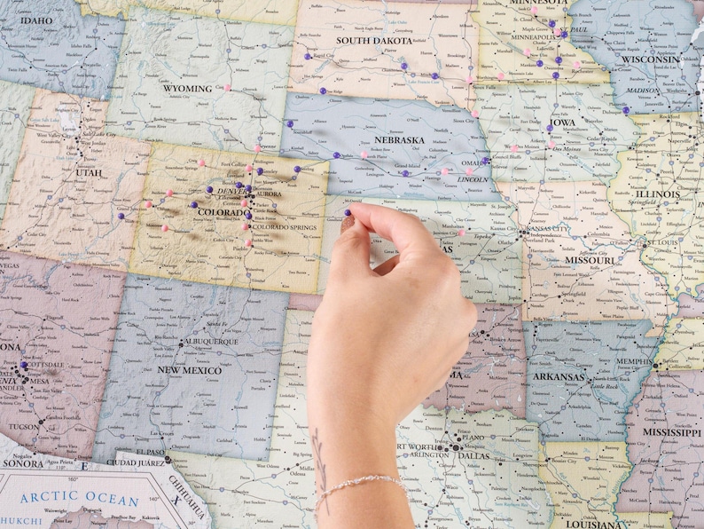 May include: A close-up of a map of the United States with push pins marking various locations. A hand is placing a push pin on the map.