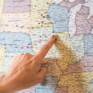 May include: A close-up of a hand pointing to the state of Illinois on a map of the United States. The map is covered in colorful push pins.