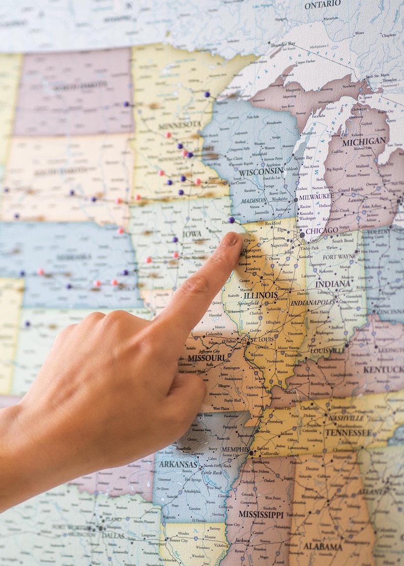 May include: A close-up of a hand pointing to the state of Illinois on a map of the United States. The map is covered in colorful push pins.