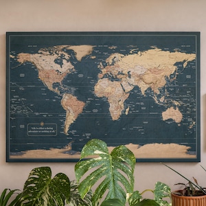 May include: A large rectangular world map wall art piece with a dark blue background and tan-coloured continents. The map features the text "Life is either a daring adventure or nothing at all." It is framed in a thin dark border and mounted on a wall.