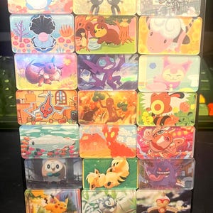 May include: A collection of seventeen rectangular acrylic magnets featuring colorful illustrations of various animated creatures in diverse settings, including forest, underwater, and urban scenes, arranged in a grid on a clear display stand.