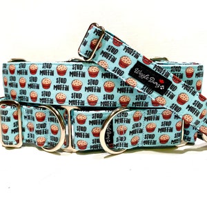May include: A light blue dog collar with a repeating pattern of red and brown cupcakes with the text "Stud Muffin" on a light blue background. The collar has silver hardware and the text "Wiggle Buns" on a red tag.