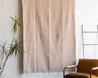 May include: A large, beige woven blanket with a fringed edge hangs on a white wall. The blanket is vertically striped. A brown leather chair with black metal legs and a plant in a black pot are in the foreground.