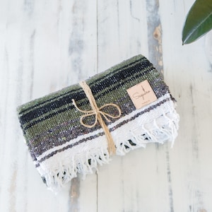 May include: A green, black, and white striped Mexican blanket with white fringe is folded and tied with twine. A brown tag with the text "Saguaro" is attached to the blanket.