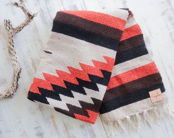 May include: A woven throw blanket with a geometric pattern in shades of red, black, and white. The blanket has a fringe border and a brown leather tag with the word "Sapien" printed on it.