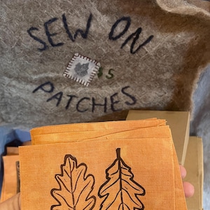 May include: A stack of orange fabric squares with a brown block print of two oak leaves. The fabric is labeled "Sew On Patches".