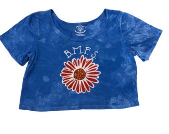 May include: A blue, hand-dyed t-shirt with a scoop neck and short sleeves. The shirt features a large, red and white daisy graphic with the letters "B.M.F.S." above it. The shirt has a tie-dye pattern.