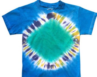 May include: A blue tie-dye t-shirt with a central green diamond shape, surrounded by a circular pattern of yellow, purple, and white. The shirt has short sleeves and a crew neck. The words "hand dyed" are printed on the top of the shirt.
