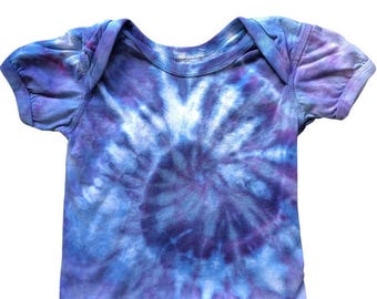 May include: A tie-dye baby bodysuit in shades of blue and purple. The short-sleeved onesie features a spiral pattern in the center, with a light blue and purple color scheme. The fabric is soft and appears to be cotton.