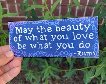 May include: A rectangular sticker with a blue background and white text that reads "May the beauty of what you love be what you do" by Rumi. The sticker has a decorative border and is held in front of green plants.