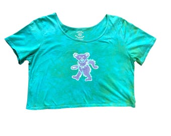 May include: A turquoise tie-dye t-shirt with a purple dancing bear graphic. The shirt has a scoop neck and short sleeves. The text "Dead Head by Karen Lee" is printed on the neckline.