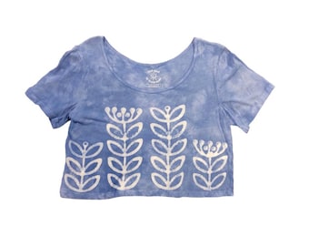May include: A light blue, short-sleeved crop top with a scoop neck. The shirt features a white floral design across the front. The fabric has a tie-dye effect. The shirt is made of a soft material.