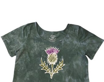 May include: A dark green tie-dye crop top with a scoop neckline and short sleeves. The center features a graphic of a thistle plant with a purple flower head and green leaves. The interior neck label reads Hand Dyed by WAXON Studio.