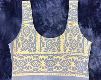 May include: A light blue crop top with a yellow, symmetrical pattern. The pattern features geometric shapes and floral elements. The top has a scoop neck and wide straps, and is made of a soft fabric.