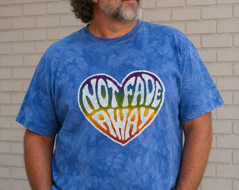 May include: A blue tie-dye t-shirt featuring a large heart-shaped graphic in the center. The heart has a rainbow-colored border and contains the white text "NOT FADE AWAY" in a stylized, wavy font. The shirt has short sleeves and a crew neckline.