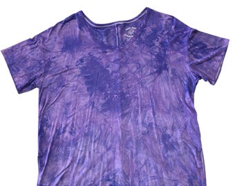 May include: A purple tie-dye t-shirt with a scoop neck. The shirt has short sleeves and a relaxed fit. The fabric has a mottled pattern of light and dark purple hues. The text "hand dyed by Watson" is visible on the neckline.