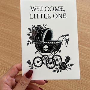 May include: A white greeting card featuring black ink illustrations of a baby carriage adorned with roses and a skull motif. The text WELCOME, LITTLE ONE is printed in black at the top. The carriage has four wheels and a bow hanging from the handle.
