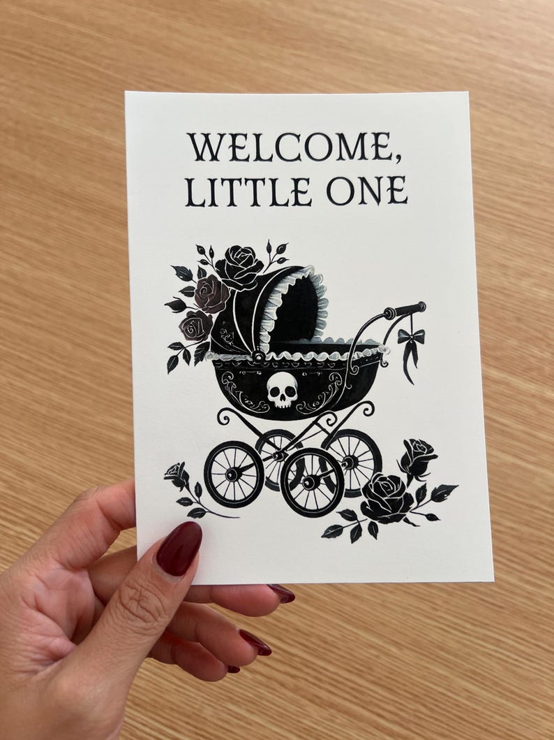 May include: A white greeting card featuring black ink illustrations of a baby carriage adorned with roses and a skull motif. The text WELCOME, LITTLE ONE is printed in black at the top. The carriage has four wheels and a bow hanging from the handle.