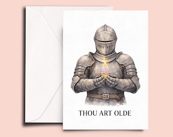 May include: Greeting card featuring a watercolor illustration of a knight in full armor holding a cupcake with a lit candle. The text "THOU ART OLDE" is printed below the knight. A white envelope is included.