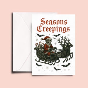 May include: A white greeting card with a matching envelope featuring a red and black illustration of a skeleton dressed as Santa Claus riding a sleigh pulled by two skeletal reindeer. The text Seasons Creepings is printed in red above the scene.