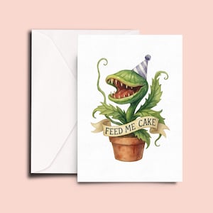 May include: Greeting card featuring a green plant with a large, toothy mouth wearing a party hat. The plant is in a brown pot, with a banner that reads "FEED ME CAKE". A white envelope is on the left.