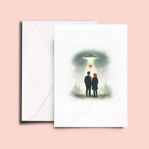May include: A greeting card featuring a watercolor illustration of two figures standing in a forest at night, looking up at a glowing UFO. A small red heart floats in the beam of light emitted by the spacecraft. A plain white envelope is included.