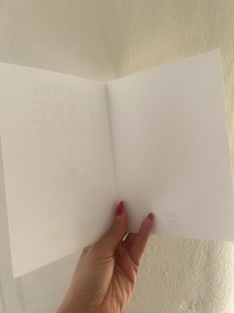 May include: A white greeting card held open to reveal a blank interior. The left side of the card features faint, partially visible text printed in a light gray font that reads MAY THE CAKE BE WITH YOU, while the right side remains completely blank.