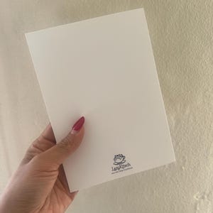 Pu&ograve; includere: Una carta rettangolare bianca tenuta in mano con smalto rosso. La carta presenta un piccolo logo nero e il testo "LazyKitsch" e un indirizzo web. Lo sfondo &egrave; un muro beige chiaro.