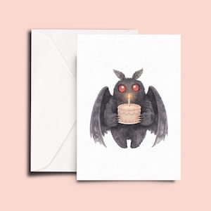 May include: A greeting card featuring a dark gray, furry moth creature with large, glowing red eyes. The creature is holding a small, light-colored cake with a single lit candle on top. The card is accompanied by a plain white envelope.